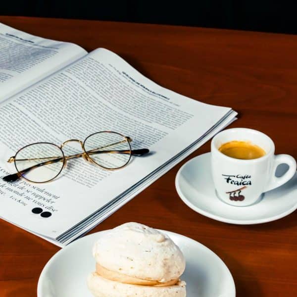 Un expresso accompagné de la pâtisseries pavlova et d'un journal superposé par des lunettes de vue.