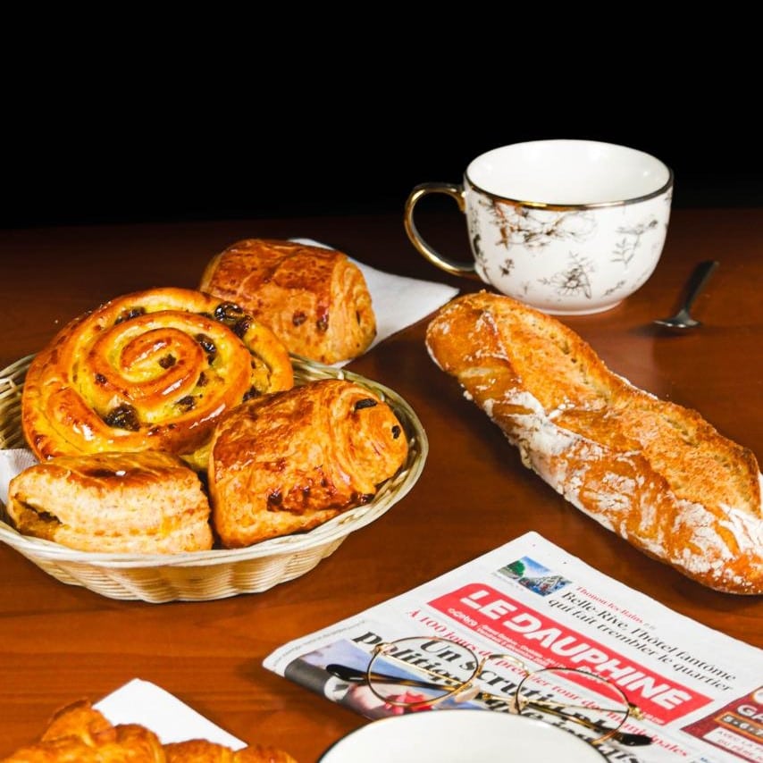 Typique petit déjeuner français montrant les viennoiseries populaires (pain au chocolat, croissant, pain au raison) avec une baguette, un journal et deux tasses ornementées.